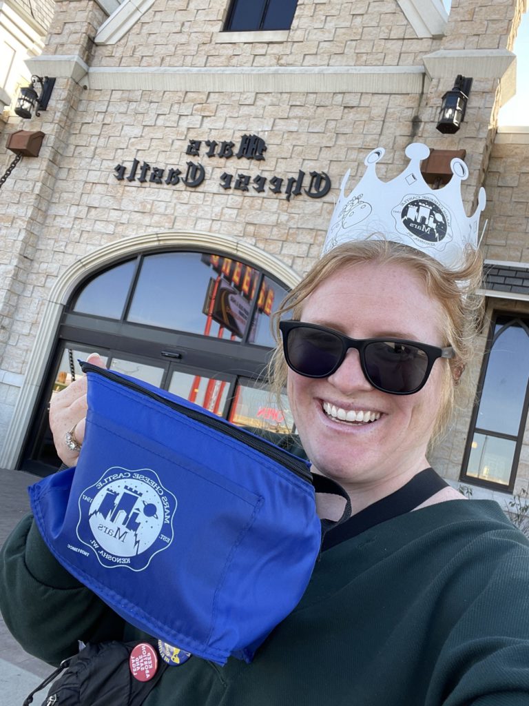 Morgen smiling wearing sunglasses and a paper crown, showing off the Mars Cheese Castle cooler bag