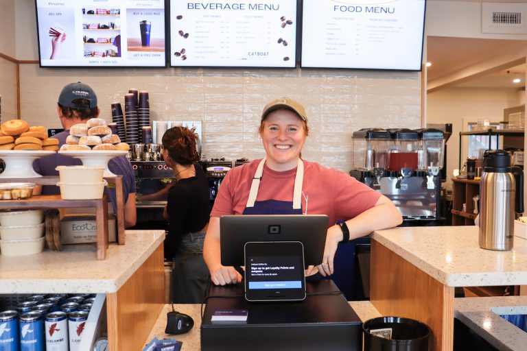 A photo of Morgen, behind the cash register at Catboat Coffee Co. Big smile and leaning on the counter with the espresso machine and coffee grinders in the background. 